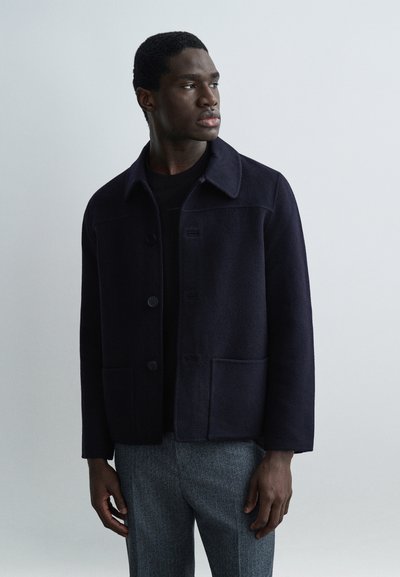 Hombre con chaqueta de lana azul marino oscuro sobre una camiseta negra y pantalones grises, de pie y mirando hacia un lado contra un fondo liso.