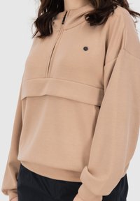 Felpa con zip beige, colletto alto, spalle scese e polsini larghi. Presenta un bottone di accento nero e un design aderente a strati in vita.