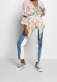 Osoba v patterned tunice s tkaničkou v pase, dvoubarevných modro-bílých džínách, olivově zelených teniskách a náhrdelníku na bílém pozadí.