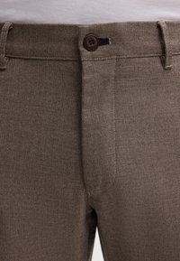 Pantaloni intrecciati marroni con una texture liscia, dotati di chiusura a bottone e passanti per cintura, mostrati all'altezza della vita e della parte superiore della coscia.