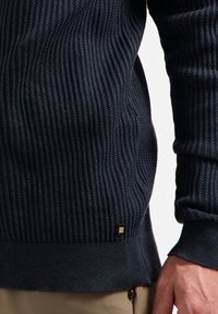 Marineblauwe, textuurtrui met geribbeld patroon, voorzien van een aansluitende manchette en onderrand. Kleine metalen accenten bij de linkeronderkant.