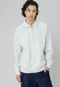 Jeune homme avec des cheveux noirs bouclés portant un sweat à capuche gris clair zippé et un jean bleu foncé, se tenant contre un arrière-plan gris uni.