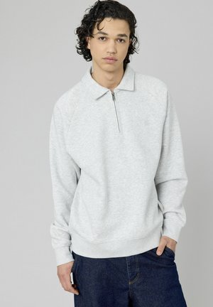 Jeune homme avec des cheveux noirs bouclés portant un sweat à capuche gris clair zippé et un jean bleu foncé, se tenant contre un arrière-plan gris uni.