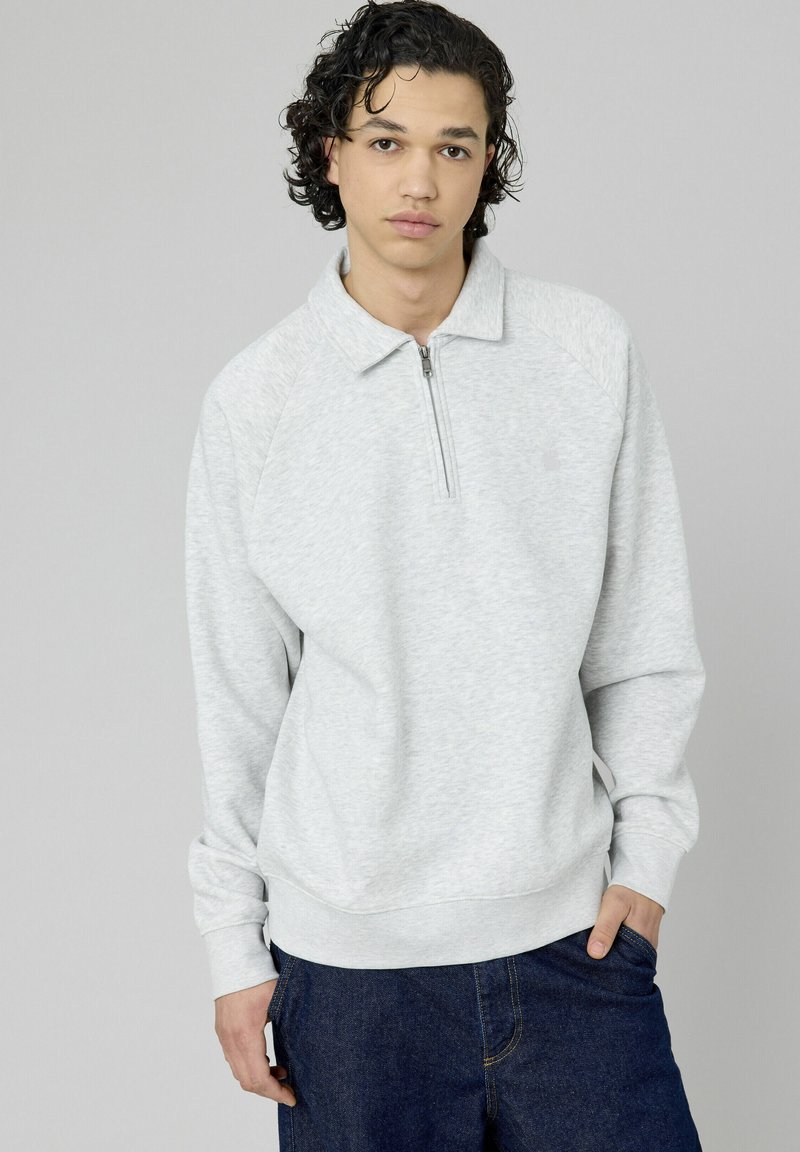 Jeune homme avec des cheveux noirs bouclés portant un sweat à capuche gris clair zippé et un jean bleu foncé, se tenant contre un arrière-plan gris uni.