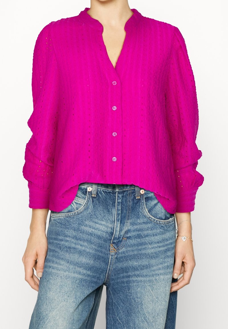 Fuchsia blouse met knopen en een textuurpatroon, met een omgeslagen kraag en volumineuze mouwen. Gecombineerd met hoog opgesneden blauwe denim jeans.