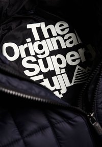 Musta, tikattu takki, jossa suuri valkoinen logo, jossa lukee "The Original Super Fuji". Sileä pinta, vetoketjusuljenta ja kauluksen yksityiskohta.