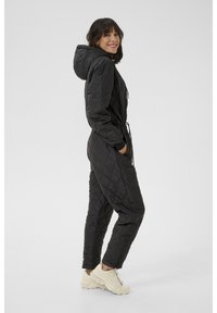 Schwarzer gesteppter Jumpsuit mit Kapuze, verstellbarem Kordelzug in der Taille, gerippten Bündchen und Seitentaschen, kombiniert mit hellen Sneakers.
