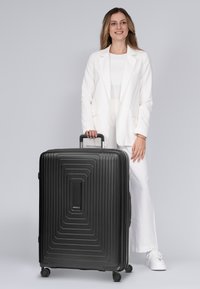 Maleta rígida negra con diseño lineal texturizado, mango lateral y ruedas giratorias. Logo en la parte frontal. Colocada al lado de una persona con atuendo claro.