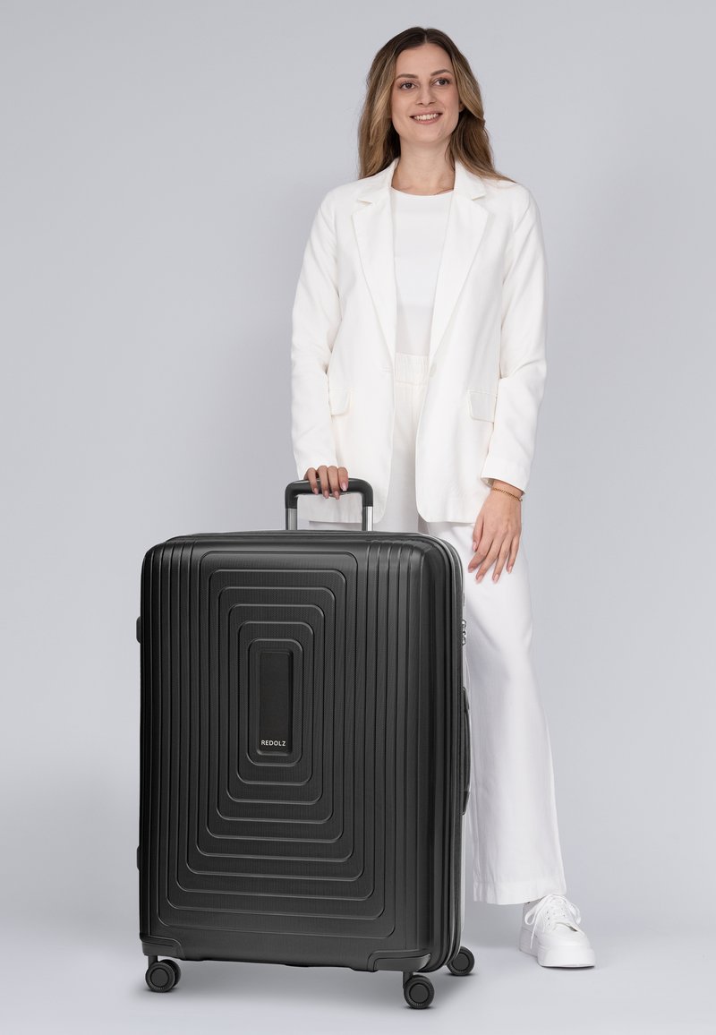 Maleta rígida negra con diseño lineal texturizado, mango lateral y ruedas giratorias. Logo en la parte frontal. Colocada al lado de una persona con atuendo claro.