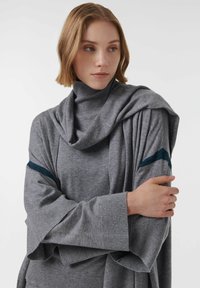Suéter de cuello alto gris con diseño drapeado, mangas sueltas y puños acanalados. Rayas teal notables en las mangas añaden contraste. Textura suave.