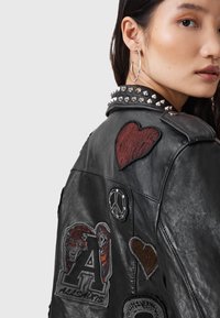 Woman wearing black leather jacket with silver spikes on collar and various embroidered patches, including a red heart and a tiger logo.