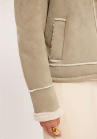 Zielona kurtka z sztucznego zamszu z kremowymi akcentami z barankowej wełny i przeszyciami, z boczną kieszenią oraz wzorzystym białym swetrem z prążkowanym wzorem pod spodem.