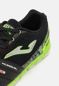 Baskets de sport noires avec accents verts, tige en mesh et motif texturé sur le côté. Présente un logo gris prononcé et une semelle en caoutchouc durable.