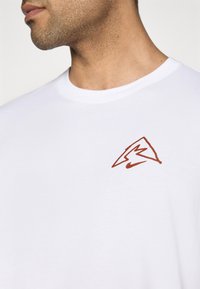 Hvit bomull t-skjorte med rund hals og korte ermer. Har en brun logodesign på venstre brystområde. Glatt tekstur.