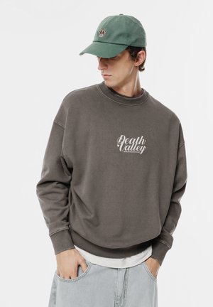 Szary sweter z napisem "Death Valley", ściągaczem przy dekolcie i luźnym krojem, nałożony na biały T-shirt. Zielona czapka z motywem misia.