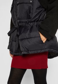 Chaqueta negra acolchada con cinturón en la cintura y mangas de forro polar, llevada sobre falda de punto roja y medias negras, con la mano colocada en el bolsillo de la chaqueta.