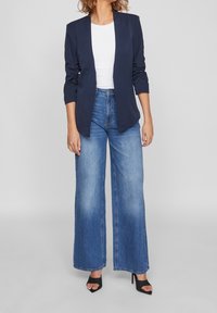 Blazer bleu marine à coupe ajustée et à manches longues, associé à un jean bleu taille haute à jambes larges et des escarpins noirs à bout ouvert.