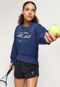 Σκούρο μπλε φούτερ με λευκή τυπωμένη μάρκα, υφασμάτινη υφή και ριγέ τελειώματα. Συνδυασμένο με μαύρες βερμούδες που έχουν ένα μικρό λογότυπο. Κρατάει μια πράσινη μπάλα τένις.