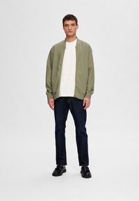 Veste bomber vert olive sur une chemise blanche, associée à un jean bleu foncé et des chaussures noires. Tenue décontractée avec des poignets et un col côtelés.