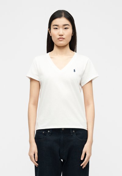 Νεαρή γυναίκα με ίσια μαύρα μαλλιά φοράει λευκό T-shirt με V λαιμόκοψη, μικρό μπλε λογότυπο και σκούρο τζιν μπροστά σε απλό φόντο.