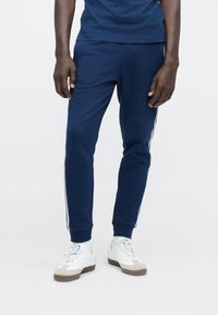 Navy zilas sporta bikses no mīksta auduma ar baltām sānu svītrām, šaurinātu piegriezumu un riboto aproču detaļām. Valkātas ar baltām kedām.