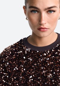 Rudas blizgučių megztinis su padažyta apykakle. Blizgučiai yra blizgūs ir skirtingų dydžių, sukuriantys tekstūruotą, atspindinčią paviršių.