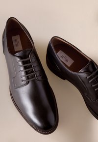 Paar polierte dunkelbraune Lederschuhe mit Schnürsenkeln auf hellbeigem Hintergrund.