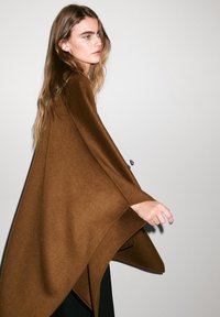 Brun ullponcho med en lös, draperad design. Har en asymmetrisk nederkant och en jämn textur, bärs över svarta kläder. Enkel halsbanddetalj.