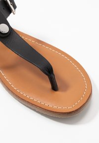 Mustat nahkaiset flip-flopit, joissa on thong-hihna, tanvärinen nahkapohja ja kontrastivärinen valkoinen ompelu reunassa.