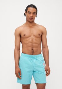 Shorts de bain turquoise avec une taille élastique et un cordon de serrage, présentant un petit logo sur la jambe gauche. Matériau lisse et léger.
