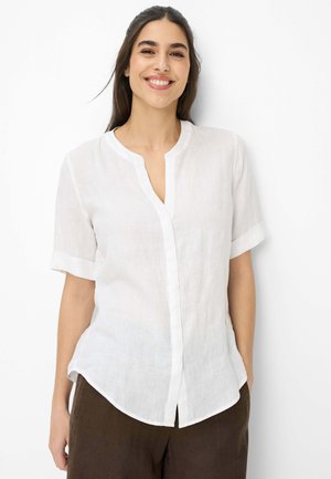 Jonge vrouw die glimlacht, draagt een wit kort mouwen blouse met knopen en een bruine broek, staand tegen een witte achtergrond.