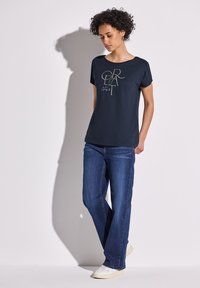 Camiseta color azul marino con cuello redondo y mangas cortas, con un ligero estampado gráfico en el centro. Combinada con unos vaqueros de corte recto de lavado claro y zapatillas blancas.