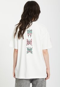 Camiseta de algodón blanca con un gráfico en la espalda que presenta tres diseños de mariposas coloridas en rosa y verde. Corte holgado con mangas cortas.