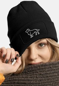 Black knit beanie with a ribbed texture and a rectangular patch featuring a white dog silhouette. Close-up of a hand holding the hat.