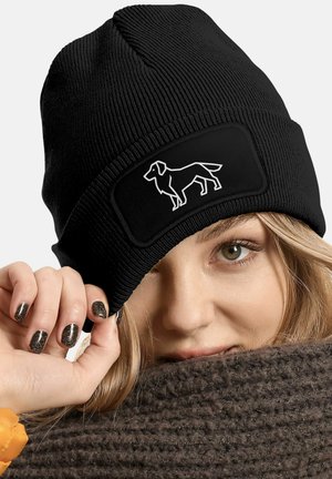 Black knit beanie with a ribbed texture and a rectangular patch featuring a white dog silhouette. Close-up of a hand holding the hat.