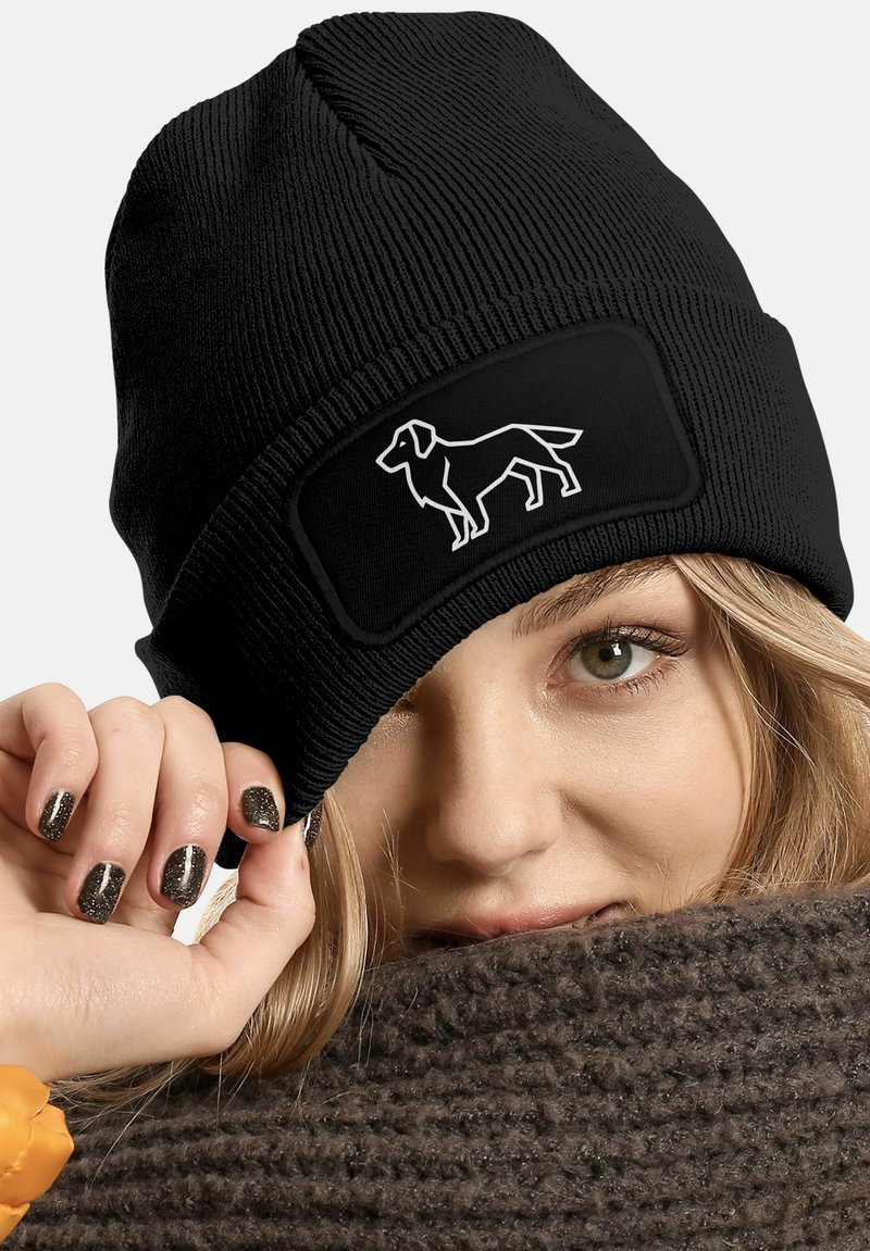 Black knit beanie with a ribbed texture and a rectangular patch featuring a white dog silhouette. Close-up of a hand holding the hat.