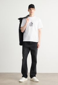 Balta T-krekls ar melnu logo, tumši denima džinsi un balti sporta apavi. Modelis tur melnu jaku pār vienu plecu, uzvelkot melnu cepuri.