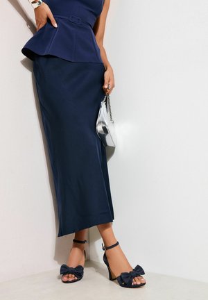 Frau in marineblauem Peplum-Oberteil und Rock, die eine silberne Handtasche hält und schwarze Sandalen mit hohen Absätzen und Schleifendetails trägt.