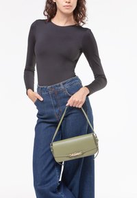 Borsa a tracolla in pelle verde oliva con forma rettangolare, caratterizzata da accessori in tono oro e una maniglia superiore, posata su pantaloni in denim a vita alta.