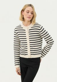 Neulecardigan, jossa on vaakasuorat mustat ja kerma-alueet, pyöreä pääntie, etutaskut ja nappikiinnitys. Pehmeä rakenne, vartalonmyötäinen malli.