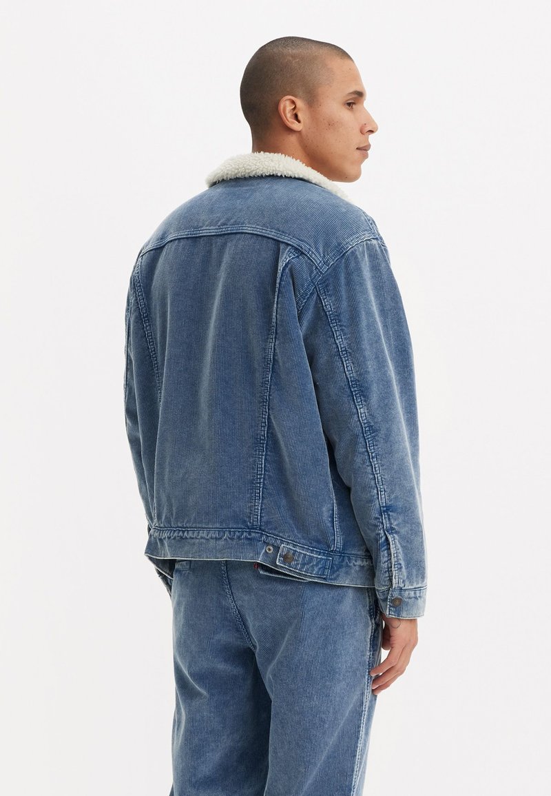 Giacca di jeans di un blu chiaro con colletto in shearling bianco. Il tessuto ha una finitura testurizzata e presenta una chiusura con bottoni e tasche laterali.