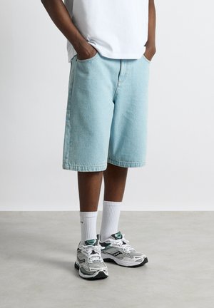 Mand iført lyseblå denim shorts til knæet, hvide crew-sokker, hvide atletik sneakers og en hvid skjorte med hænderne i lommerne.