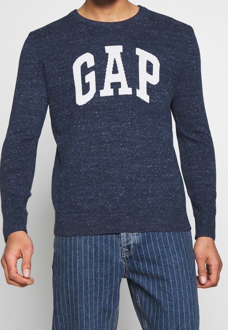 Personne portant un pull GAP bleu marine avec lettrage blanc du logo et un jean bleu à fines rayures, vue des épaules aux cuisses.