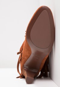 Brązowy zamszowy botek na kostkę z grubym obcasem, frędzlami po bokach i gładką podeszwą oznaczoną rozmiarem 37, oparty o białą ścianę.