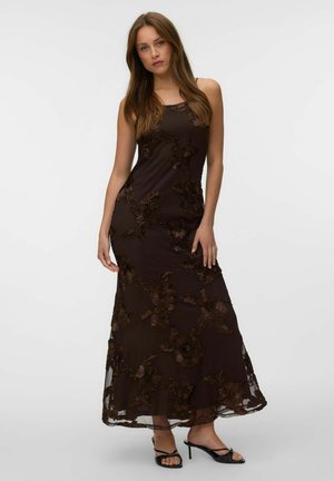 Jeune femme portant une robe sans manches marron foncé jusqu'aux chevilles avec broderie florale et des sandales noires à talons sur un fond uni.