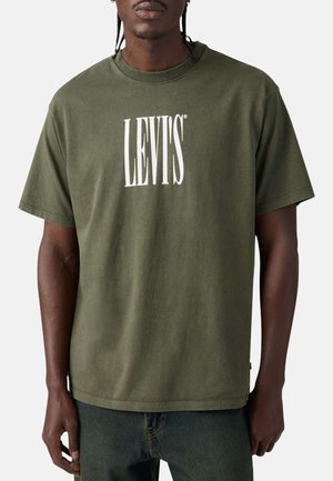 Hombre con una camiseta olive verde de Levi's con texto de logo blanco, combinada con jeans oscuros, frente a un fondo claro y liso.