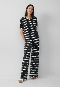 Zwart-witte stippenoverall met korte mouwen, een ontspannen pasvorm en geplooide textuur, gecombineerd met lichte sandalen.