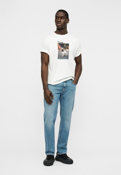 Λευκό βαμβακερό μπλουζάκι με γυρισμένα μανίκια και γραφική εκτύπωση στο κέντρο μπροστά. Συνδυασμένο με ανοιχτό μπλε τζιν παντελόνι και μαύρα αθλητικά παπούτσια.