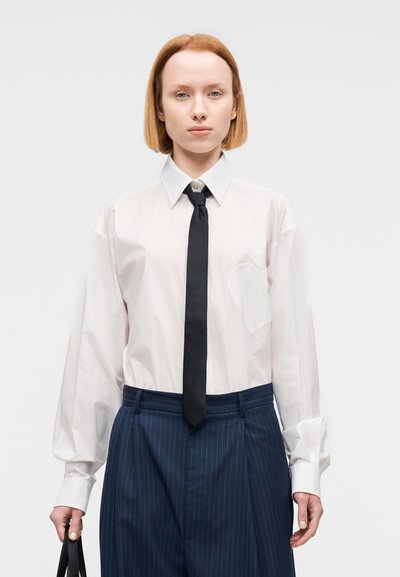 Chemise blanche boutonnée avec une cravate noire. Comprend une poche avec un accent en forme de cœur. Pantalon bleu marine à fines rayures. Le mannequin tient un sac noir.