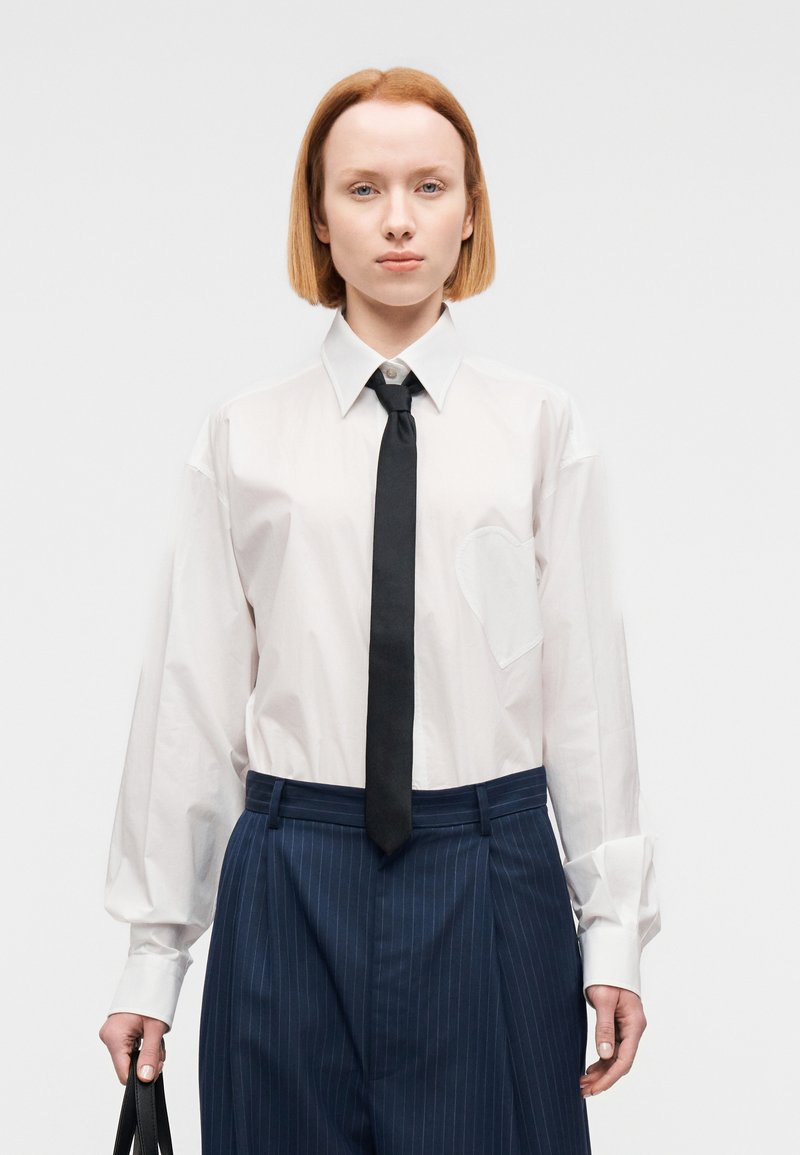 Λευκό πουκάμισο με κουμπιά και μαύρη γραβάτα. Διαθέτει τσέπη με καρδιοειδές διακοσμητικό. Μπλε σκούρο παντελόνι με λεπτές ρίγες. Το μοντέλο κρατάει μια μαύρη τσάντα.